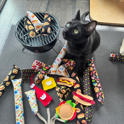Black cat near catnip toys with burgers, hot dogs, ketchup bottles, mustard bottles, and grilling inspired foodie toys