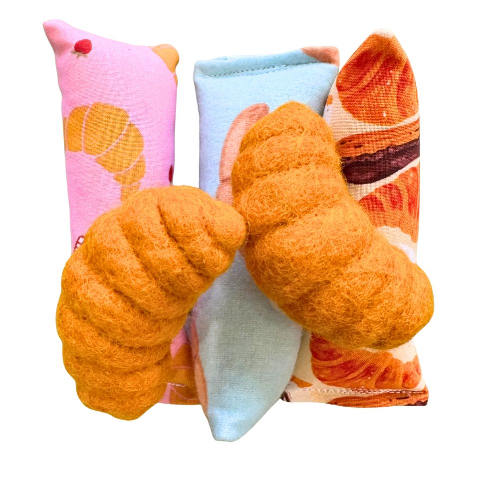 Two croissant-shaped cat toys displayed in front of three CatnipTakeout kicker catnip toys, featuring pink and blue croissant prints and a bakery-inspired pastry fabric, designed for long-lasting interactive and self-play.