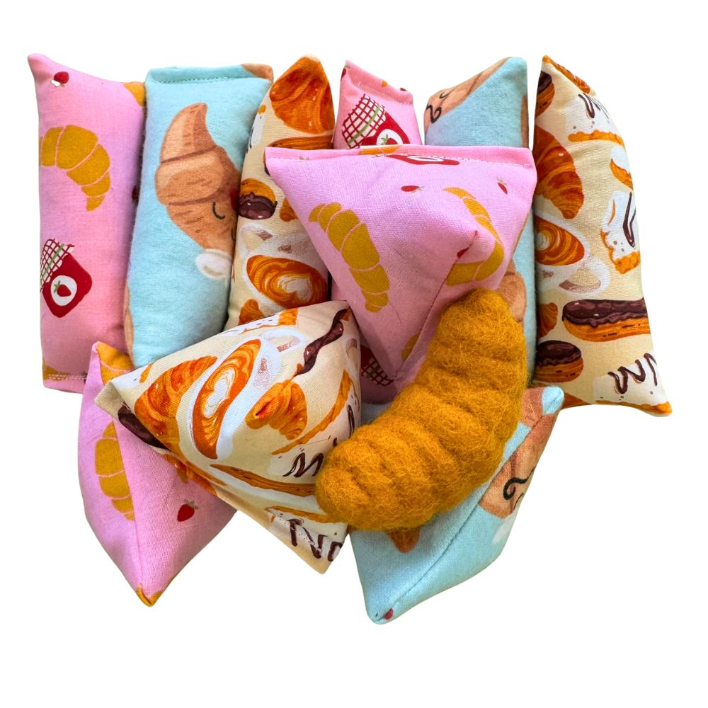 Croissant-themed cat toys arranged together, including Happy Sacks, kickers, and mini kickers made with pastel pink, blue, and cream cotton fabrics printed with croissants, café pastries, jam jars, and illustrated croissant characters, paired with a croissant-shaped felt toy, designed for durable play, batting, and bunny-kicking.