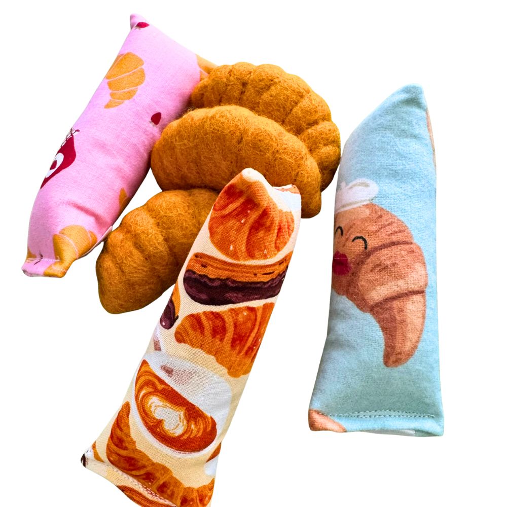 Croissant-shaped cat toys displayed with CatnipTakeout kicker and Happy Sack catnip toys, featuring pink, blue, and bakery-themed croissant fabrics, designed for handmade quality play, batting, and bunny-kicking.