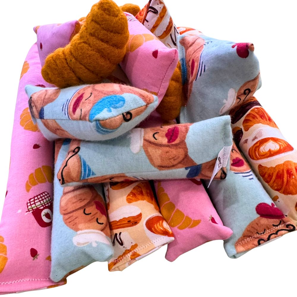 Pile of croissant-themed cat toys including Happy Sacks, kickers, and mini kickers made with pastel pink, blue, and cream cotton fabrics printed with illustrated croissants, café pastries, jam jars, and character faces, layered with a croissant-shaped felt toy, designed for handmade play, batting, and bunny-kicking.