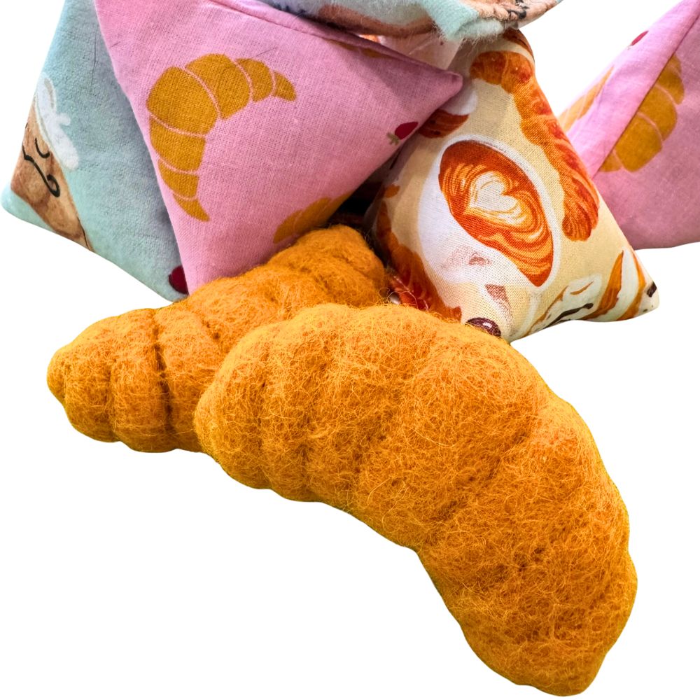Two croissant-shaped cat toys positioned in the foreground with CatnipTakeout Happy Sack catnip toys behind them, featuring pink and neutral croissant and bakery-themed fabric prints on a white background.