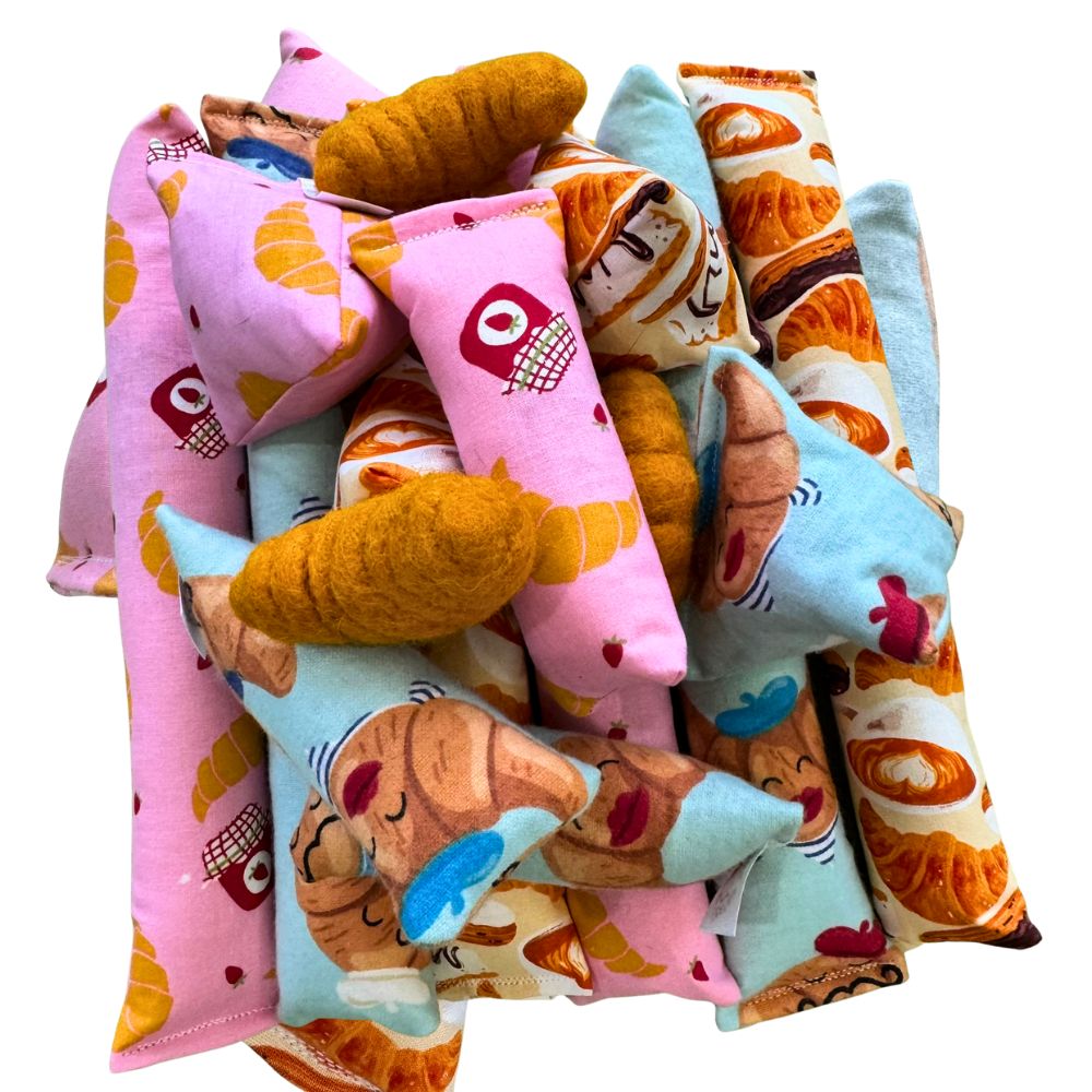 Assorted croissant-themed cat toys piled together, including Happy Sacks, kickers, and mini kickers made with pastel pink, blue, and cream cotton fabrics printed with croissants, café pastries, jam jars, and illustrated croissant characters, mixed with croissant-shaped felt toys for interactive batting and kicking play.