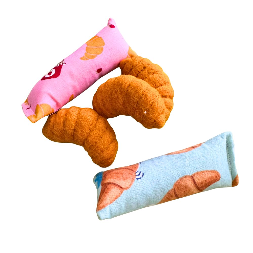 Croissant-shaped cat toys grouped with CatnipTakeout kicker catnip toys in pink and blue croissant-themed fabrics, designed for independent self-play, batting, and bunny-kickin