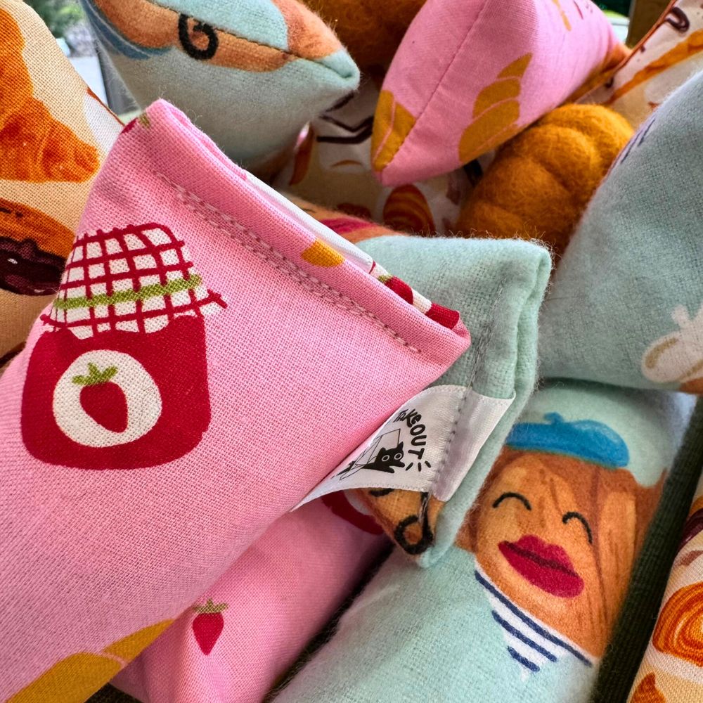 Close-up of CatnipTakeout croissant-shaped catnip toys showing reinforced stitching and brand tag, made with pastel pink and blue printed fabric featuring croissants, jam jars, and smiling pastry illustrations, designed for interactive play, batting, and kicking for kittens and cats.