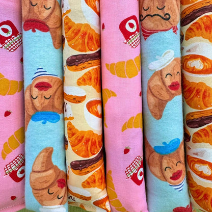 Close-up detail of CatnipTakeout Croissant Kicker catnip toys arranged tightly side by side, showing long, slim kicker shapes designed for full-body bunny kicking. The fabric features a playful bakery theme with golden croissants, chocolate pastries, pink jam jars, strawberries, and illustrated croissant characters wearing berets and chef hats. Colors include soft pink, cream, and light blue cotton fabrics with visible stitched seams. These interactive kicker cat toys are filled with CatnipTakeout’s signatu