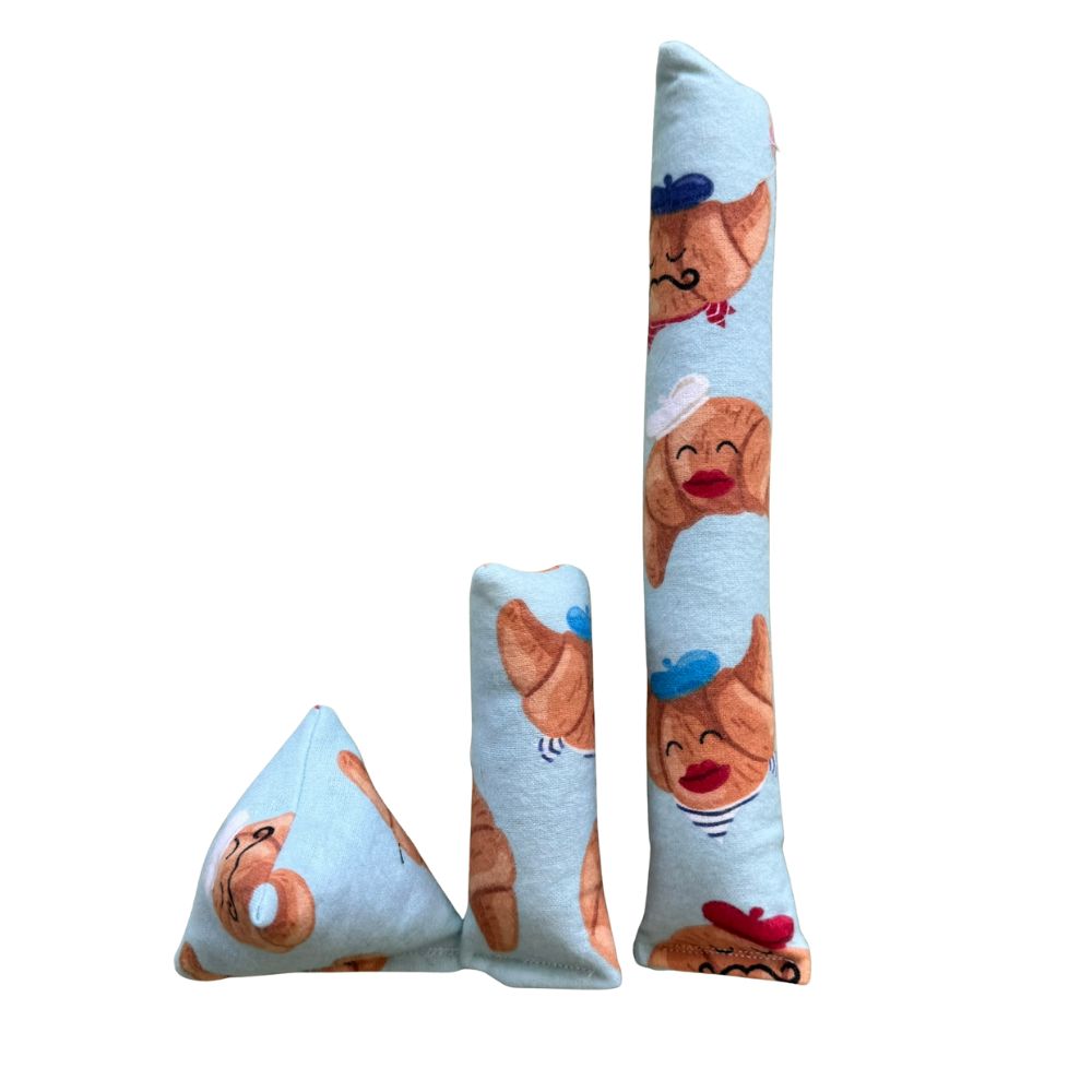 Croissant-themed cat toys shown together, including a pastel blue Happy Sack, mini kicker, and full-size kicker made with smooth cotton fabric printed with cartoon croissants wearing berets and red lips, designed for interactive batting and bunny-kicking.