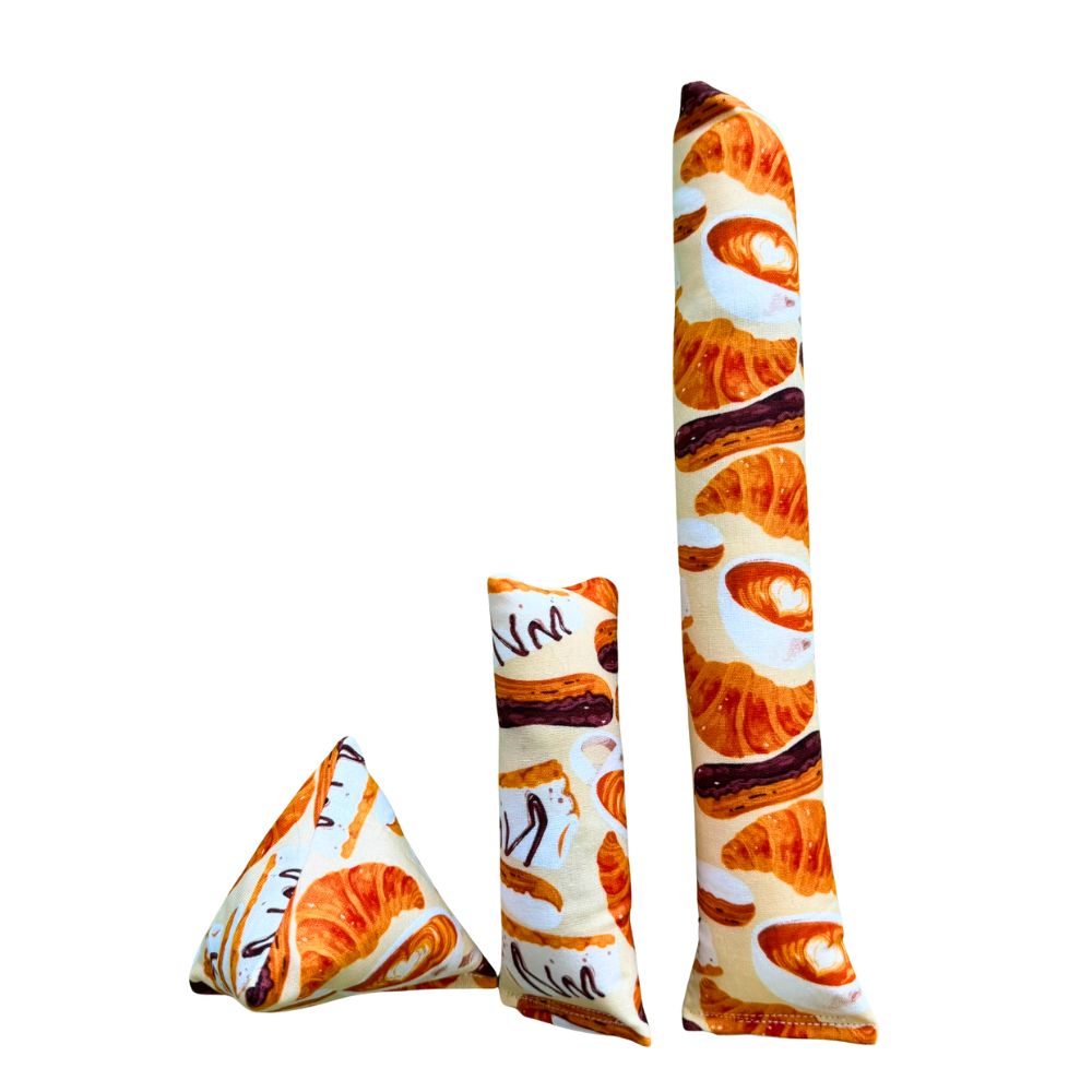 Croissant-themed cat toys shown together, including a Happy Sack, mini kicker, and full-size kicker made with cream cotton fabric printed with illustrated croissants, pastries, and café-style lettering, designed for durable play, kicking, and batting