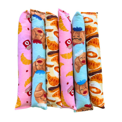 CatnipTakeout Croissant Kicker Catnip Toy lineup displayed upright in a neat row of long, slim kicker toys made for full-body bunny kicks and wrestling. The bakery-themed fabric designs include repeating golden croissants, chocolate-filled pastries, pink jam jars, strawberries, and playful illustrated croissant characters wearing berets and chef hats. Fabrics appear in soft pink, cream, and light blue tones with visible stitched seams. These kickers are filled with CatnipTakeout’s signature aromatic catnip 