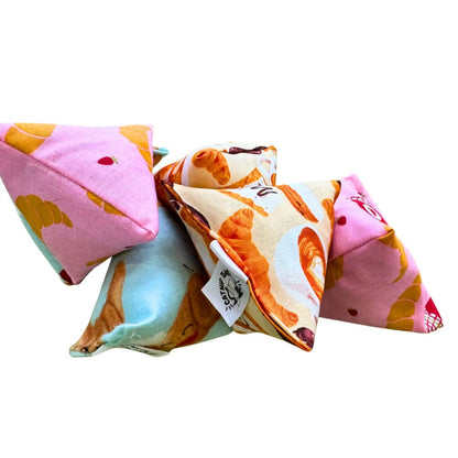 CatnipTakeout Croissant Happy Sack cat toy with a triangular pyramid design wrapped in a pink croissant bakery print fabric showing golden croissants, jam jars, and pastry accents, filled for self-play and enrichment for kittens and adult cats