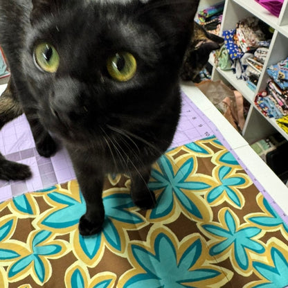 Ele Ele, a black cat with yellow-green eyes, standing on Maui Vibes Island Sand retro plumeria fabric in a CatnipTakeout workspace.
