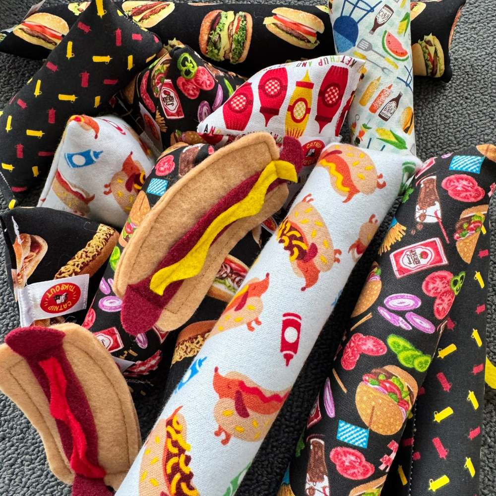 Felt hot dog cat toys beside catnip kickers with burgers, ketchup bottles, mustard bottles, and grilling inspired foodie themes