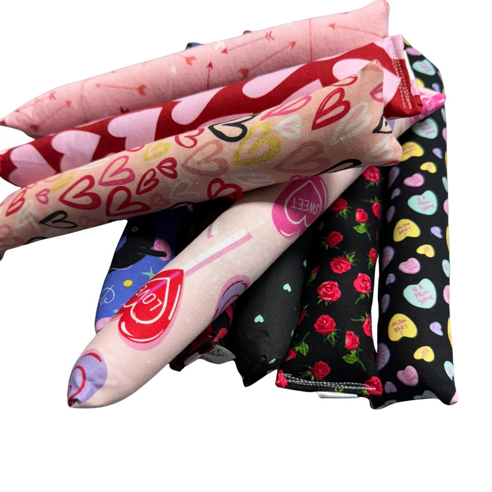 Close-up group photo of handmade Valentine cat toys showing kickers and Happy Sacks in heart prints, arrow patterns, roses, candy hearts, and black cat designs, crafted for interactive play and filled with signature premium catnip. Made by CatnipTakeout.