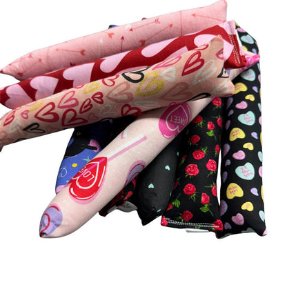 Close-up group photo of handmade Valentine cat toys showing kickers and Happy Sacks in heart prints, arrow patterns, roses, candy hearts, and black cat designs, crafted for interactive play and filled with signature premium catnip. Made by CatnipTakeout.