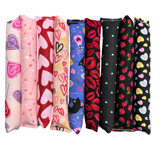 Row of handmade Valentine kicker cat toys featuring heart patterns, arrow designs, roses, lips, candy hearts, and black cat motifs, long and plush for kicking and wrestling, filled with signature premium catnip. Made by CatnipTakeout.