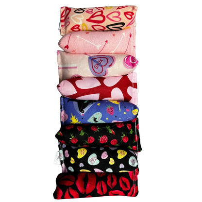 Stacked group of handmade Valentine Happy Sack cat toys in mixed designs including arrows, hearts, roses, candy hearts, and playful cats, all pyramid-shaped and filled with signature premium catnip. Made by CatnipTakeout.