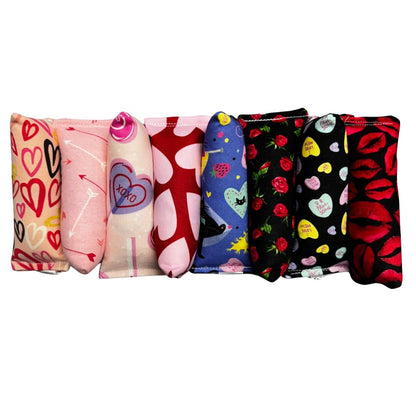 Row of handmade Valentine kicker cat toys in mixed designs including arrows, hearts, roses, candy hearts, playful cats, and bold lip patterns, filled with signature premium catnip. Made by CatnipTakeout.
